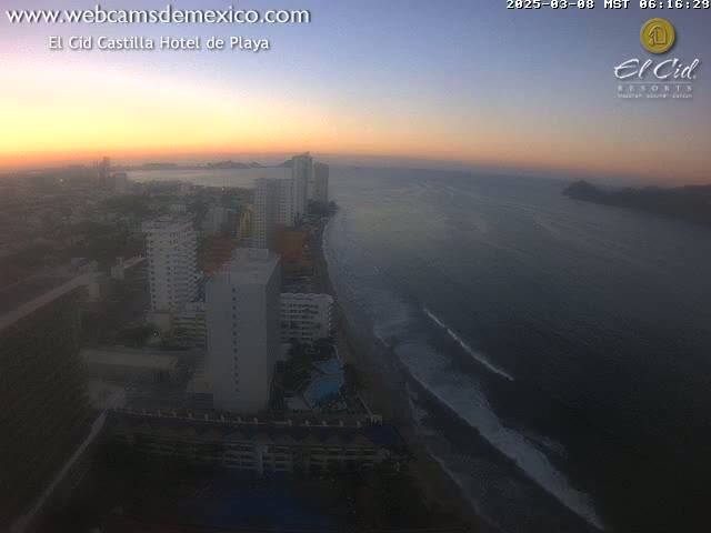 An aerial view shows a coastal city at sunset with gentle waves lapping the shore.