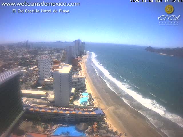 A high-angle, long shot shows a beach with a dark-sand shoreline and gentle waves lapping on the shore under a clear, sunny sky; hotels and buildings are visible inland.