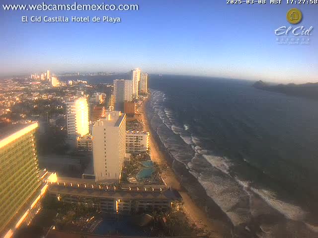 A coastal city with high-rise hotels and buildings stretches along a sandy beach with gentle waves under a clear, bright sky.