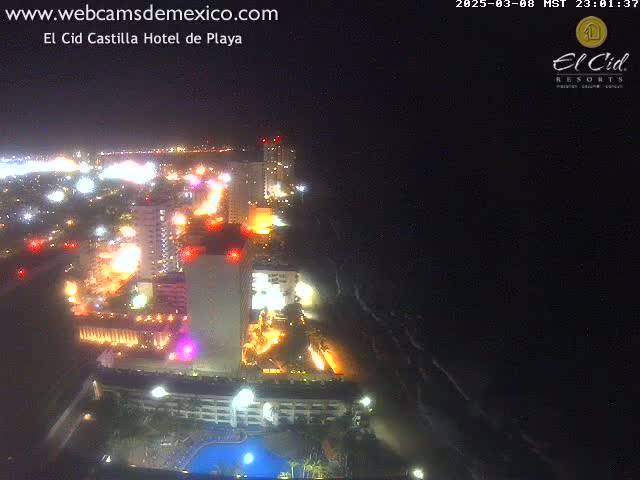 A nighttime aerial view of a brightly lit coastal city with hotels and a swimming pool, overlooking a dark ocean.