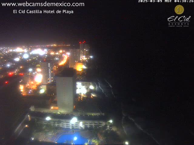 A nighttime aerial view shows a brightly lit coastal city with a hotel and swimming pool, next to dark ocean waves under a clear, dark sky.