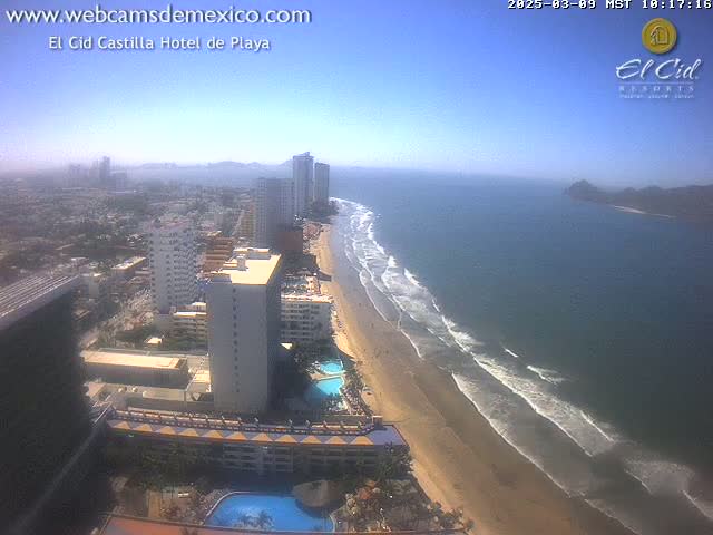 An aerial view shows a sandy beach with gentle waves lapping the shore, next to a city with high-rise buildings under a clear, sunny sky.