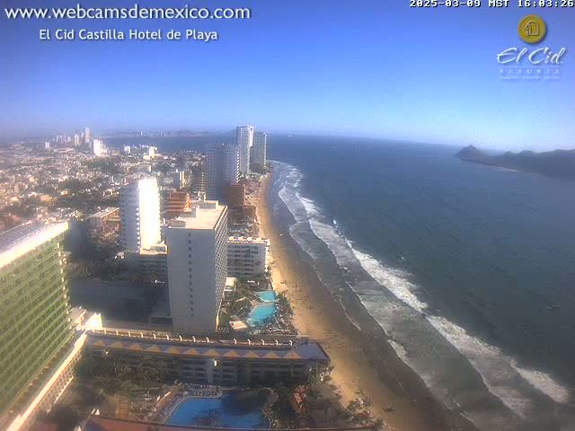 An aerial view shows a beach with gentle waves under a clear blue sky, lined with hotels and high-rise buildings in a coastal city.