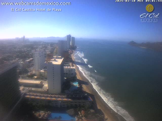 An aerial view shows a beach with small waves, high-rise hotels, and a city extending into the distance under a clear, sunny sky.