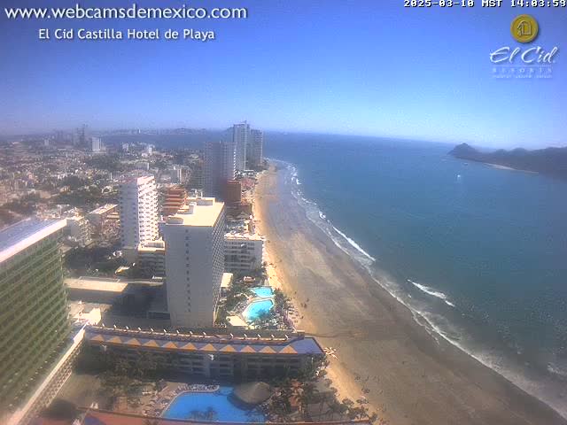 An aerial view shows a sandy beach and calm ocean next to a city with high-rise hotels under a clear, sunny sky.