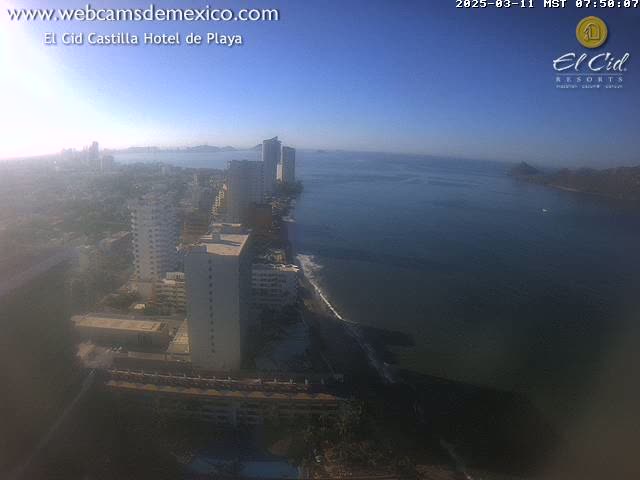 A high-angle, long shot shows a coastal city on a clear, sunny day with calm ocean waters, numerous high-rise buildings, and a dark sandy beach.
