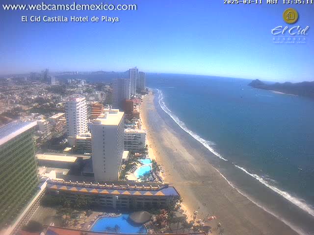 An aerial view shows a long sandy beach meeting a calm ocean under a clear blue sky, with numerous high-rise hotels and buildings lining the shore.