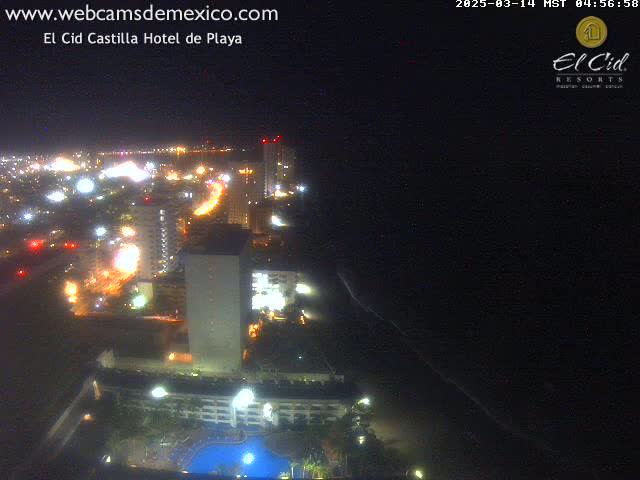 A nighttime aerial view of a coastal city, showing brightly lit buildings, roads, and a dark ocean.