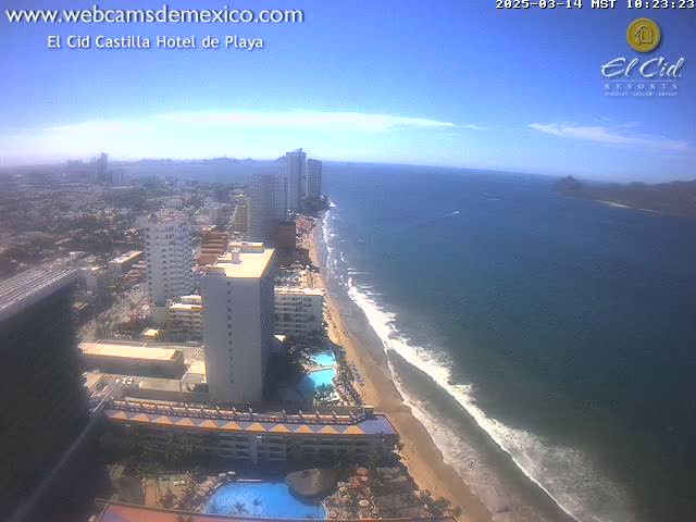 A high-angle, sunny view shows a sandy beach with gentle waves meeting a dark blue ocean, next to a cityscape of high-rise hotels and buildings.