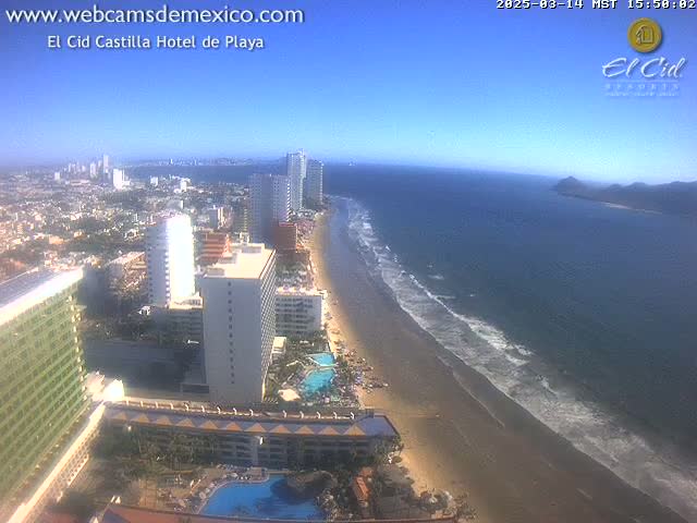 A high-angle, sunny view shows a long sandy beach with gentle waves lapping the shore, adjacent to a city with numerous high-rise hotels and buildings.