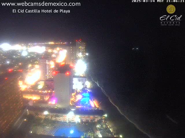 A brightly lit beachfront hotel and surrounding area are visible at night, with dark ocean visible in the foreground.