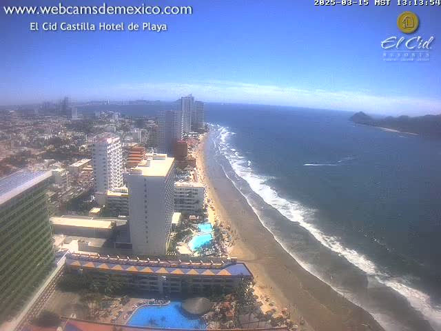 A high-angle, sunny view of a beach with calm waves, lined with hotels and high-rise buildings, extending into a city.