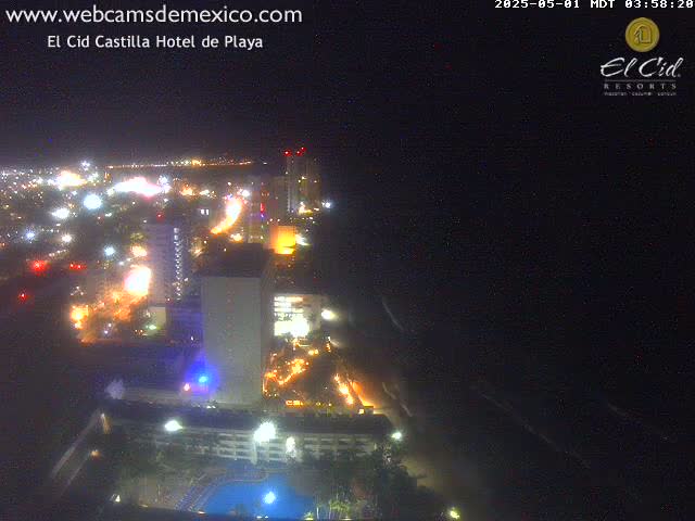 A nighttime aerial view of a brightly lit coastal city with a dark ocean and calm waves.