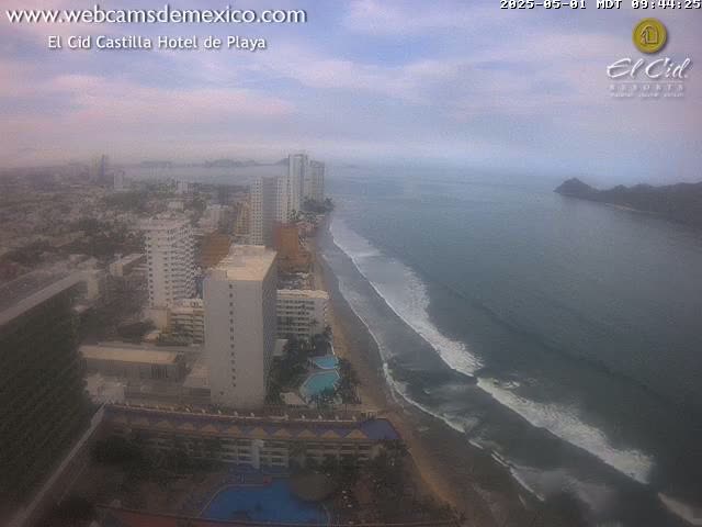 A hazy coastal city scene shows a long sandy beach with waves breaking on the shore, high-rise hotels, and other buildings stretching inland under a partly cloudy sky.
