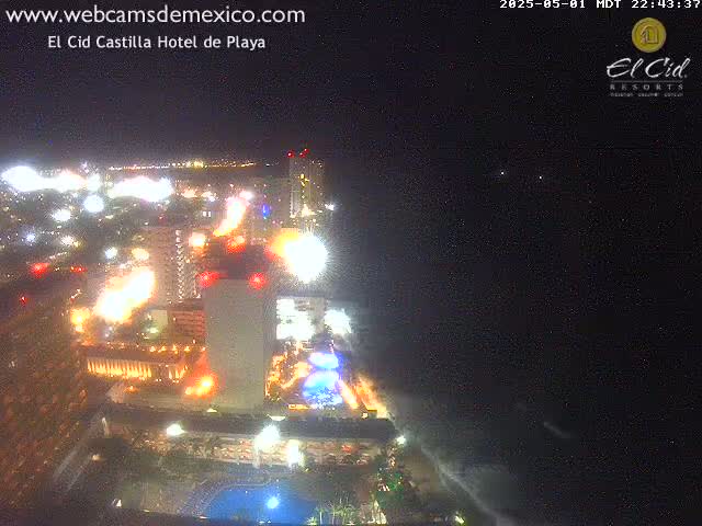 A brightly lit hotel complex at night, next to a dark ocean under a clear, starry sky.