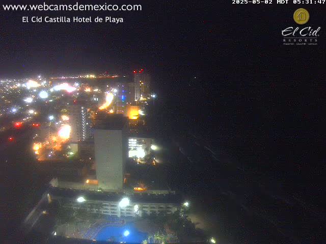 A nighttime, high-angle view shows a brightly lit city coastline with a dark ocean and a hotel complex with a pool.