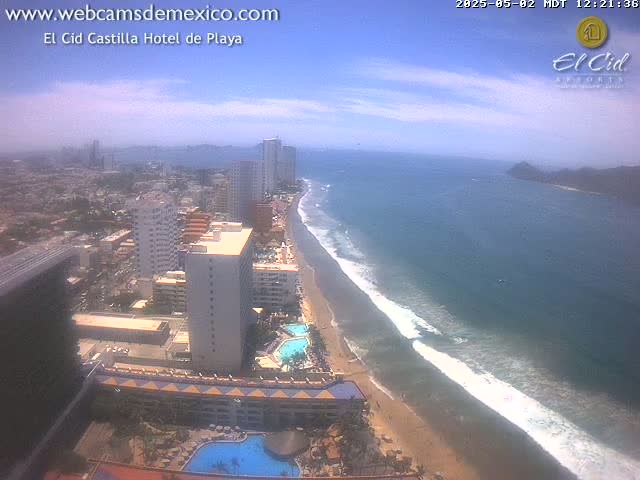 An aerial view shows a beach with waves lapping the shore, beside a city with high-rise hotels under a mostly sunny sky.