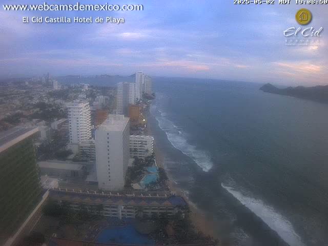 An aerial view of a coastal city at dusk, showing hotels, buildings, a beach with waves, and calm ocean water under a partly cloudy sky.