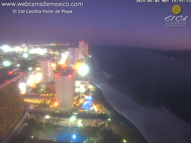 A nighttime, high-angle view shows a brightly lit resort area on a beach next to a dark ocean under a twilight sky.