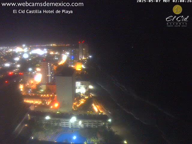 A nighttime, aerial view of a brightly lit coastal city with a dark ocean showing gentle waves.