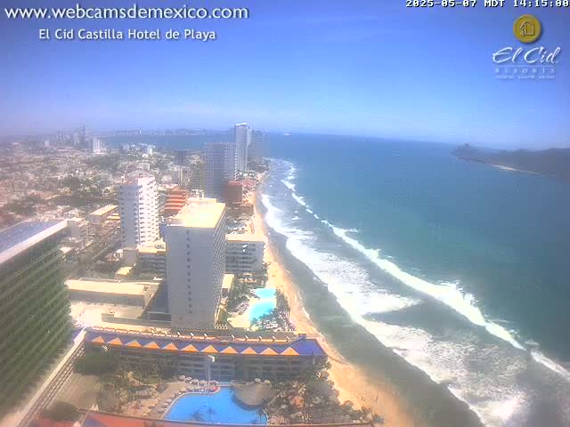 An aerial view shows a sunny beach with waves rolling onto the shore, lined with high-rise hotels and buildings, extending to a city in the background.