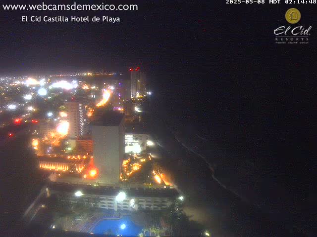 A brightly lit coastal city at night, viewed from above, shows a dark ocean with waves lapping the shore.