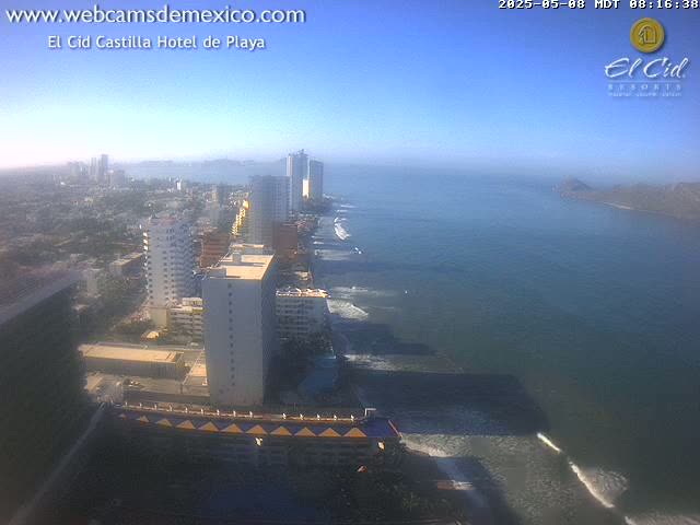 A coastal city with high-rise buildings stretches along a sandy beach and dark-blue ocean under a clear, sunny sky.