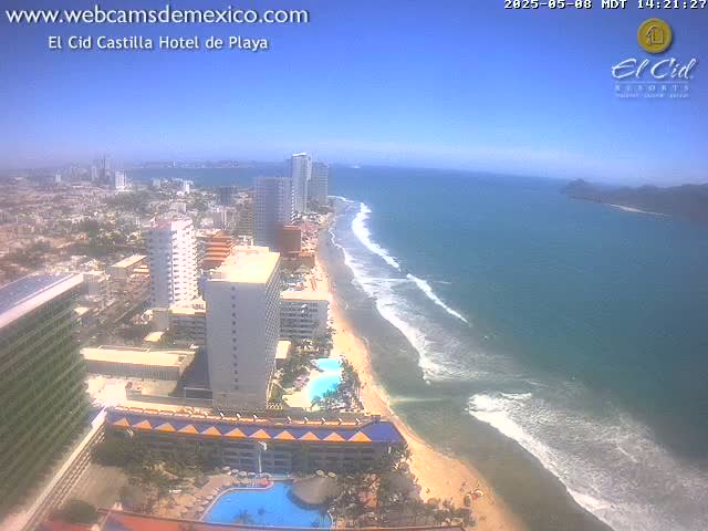 An aerial view shows a beach with light-colored sand and gentle waves under a clear, sunny sky, with high-rise hotels and buildings along the coast.