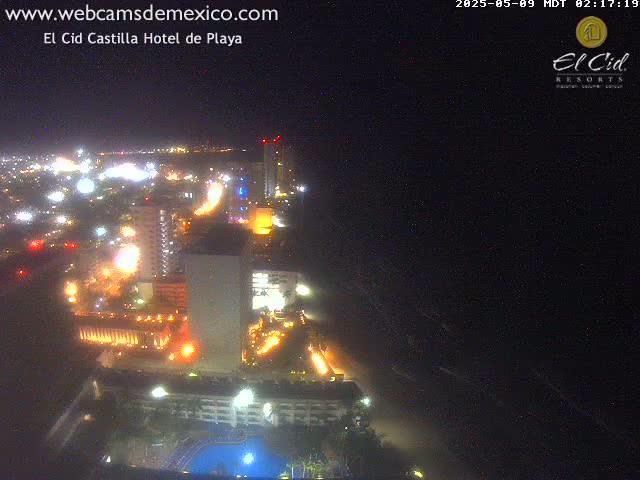 A nighttime aerial view of a brightly lit coastal city with a large hotel complex featuring a swimming pool, under a clear, dark sky.