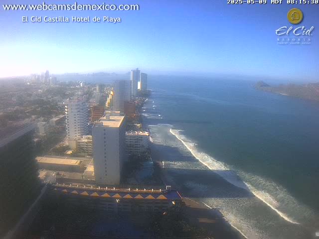 A high-angle, long shot shows a coastal city on a sunny day with ocean waves rolling onto a sandy beach.