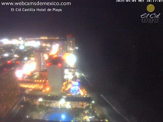 A nighttime aerial view shows a brightly lit hotel complex and surrounding city lights near a dark ocean under a clear sky.