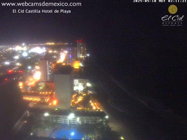 A nighttime aerial view shows brightly lit hotels and city streets along a dark ocean shoreline.