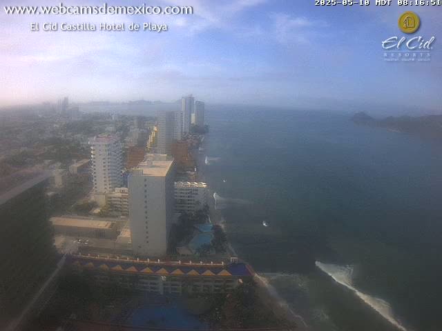 A hazy, high-angle view shows a coastline with numerous high-rise buildings and a calm ocean under a mostly clear sky.