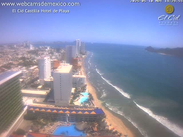 An aerial view of a beach town on a sunny day, showing hotels, buildings, and a sandy beach with waves lapping the shore.