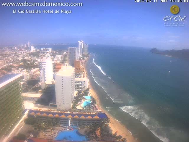 An aerial view shows a sunny beach with ocean waves lapping the shore, lined with hotels and high-rise buildings, extending to a city in the background.