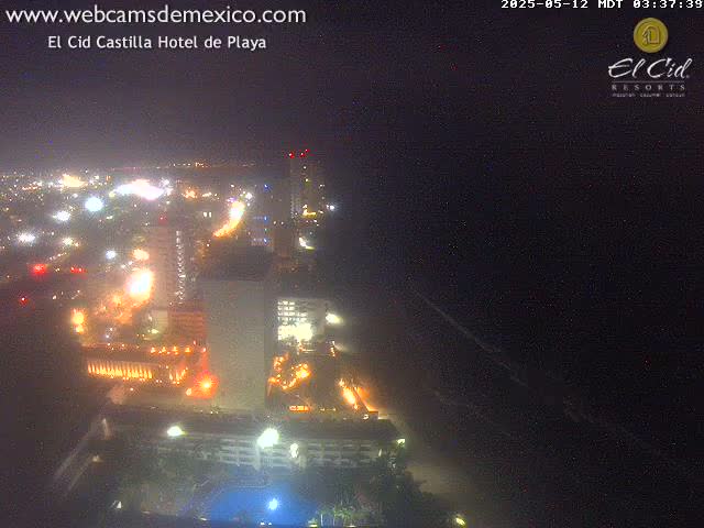 A nighttime, high-angle view shows brightly lit hotels and city lights along a dark beach under a clear sky.