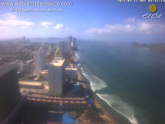 A mostly sunny coastal city scene shows high-rise hotels and buildings along a sandy beach with gently rolling waves on a partly cloudy day.