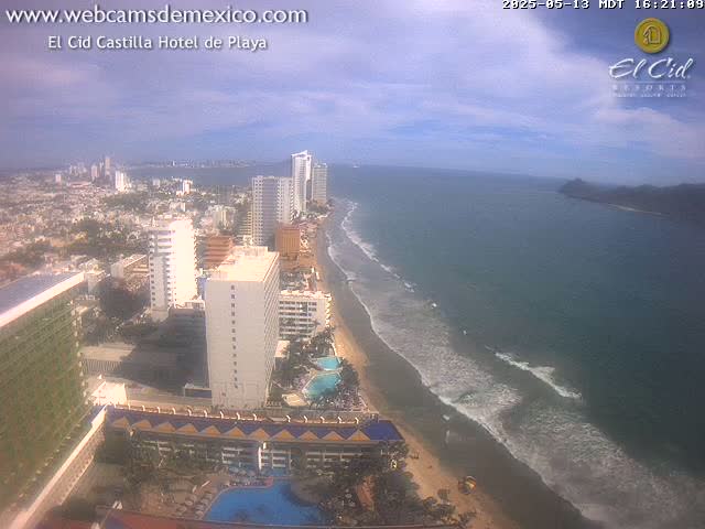 An aerial view shows a beach with gentle waves, a coastal city with high-rise hotels, and a partly cloudy sky.
