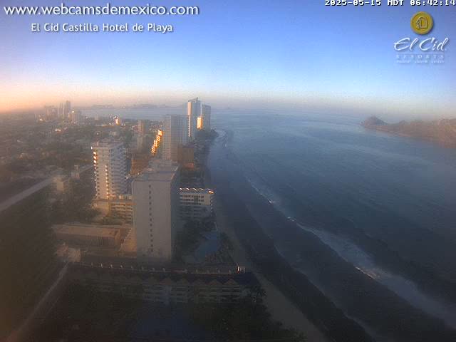A coastal city at sunrise, showing high-rise hotels and a dark sandy beach with gentle waves on a calm ocean.