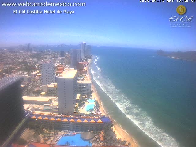 A high-angle view shows a long sandy beach with gentle waves lapping the shore, next to a city with many high-rise buildings under a clear, sunny sky.