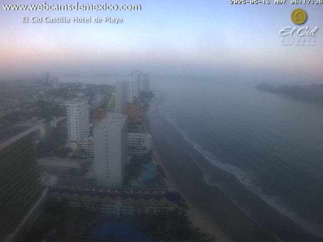 A hazy, coastal view shows a beach with gentle waves, numerous high-rise buildings, and a calm ocean extending to distant hills.