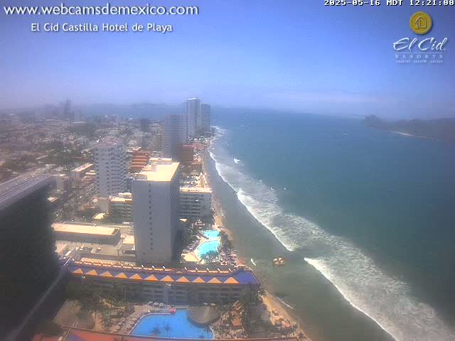 An aerial view shows a beach with gentle waves under a clear blue sky, and a coastal city with high-rise hotels and buildings stretching into the distance.