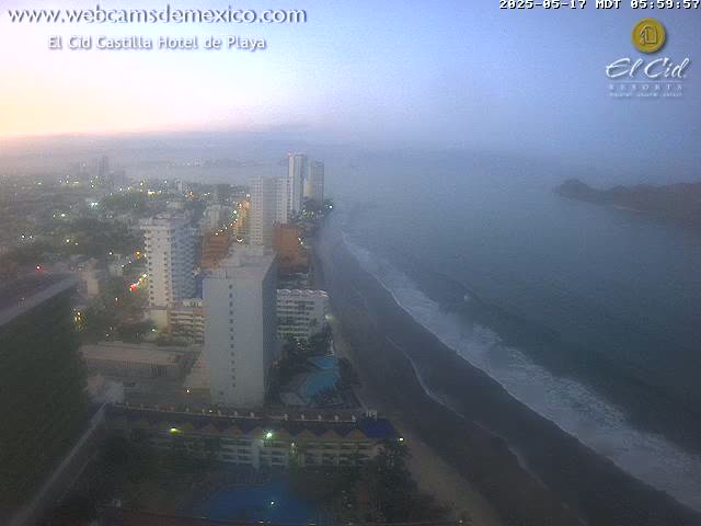 A hazy sunrise illuminates a coastal city with high-rise hotels and a dark-sand beach, where gentle waves lap the shore.