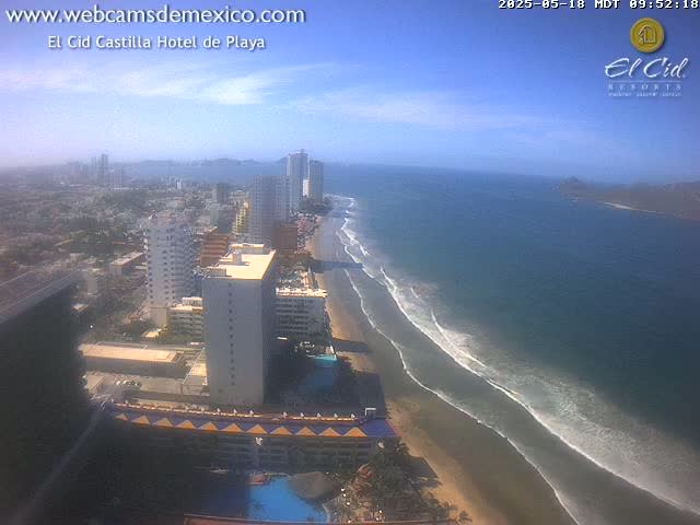 A high-angle view of a beach with gentle waves under a mostly clear sky shows a coastal city with high-rise buildings.
