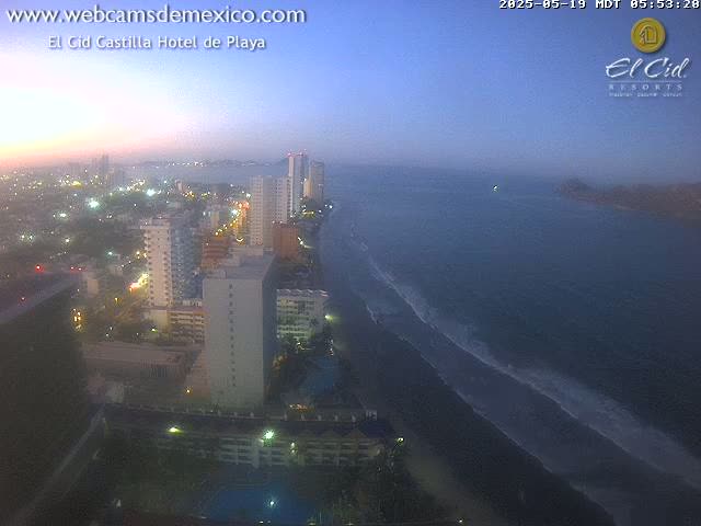 A twilight view shows a city's high-rise buildings along a dark-sand beach with gentle waves rolling onto the shore under a dusky sky.