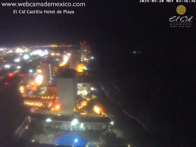 A nighttime aerial view shows brightly lit buildings along a coastline, with dark ocean waves visible below.
