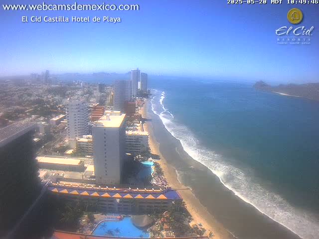 An aerial view shows a beach with gentle waves on a clear, sunny day, next to a city with high-rise hotels and buildings.