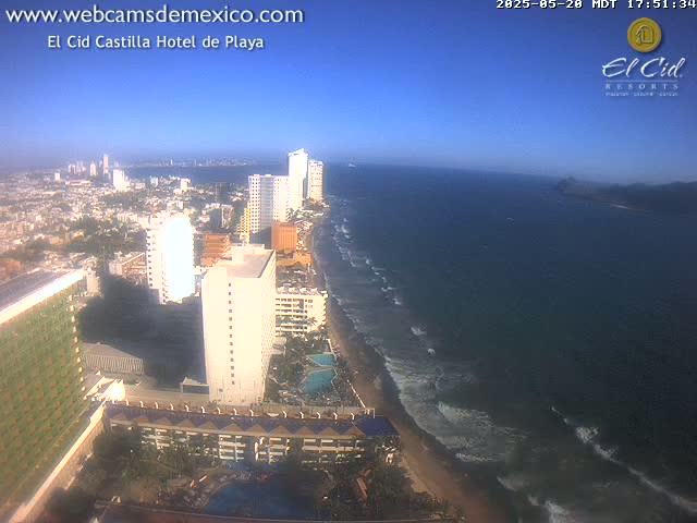 An aerial view shows a beach with waves lapping the shore, numerous high-rise hotels and buildings along the coastline, and a clear blue sky.