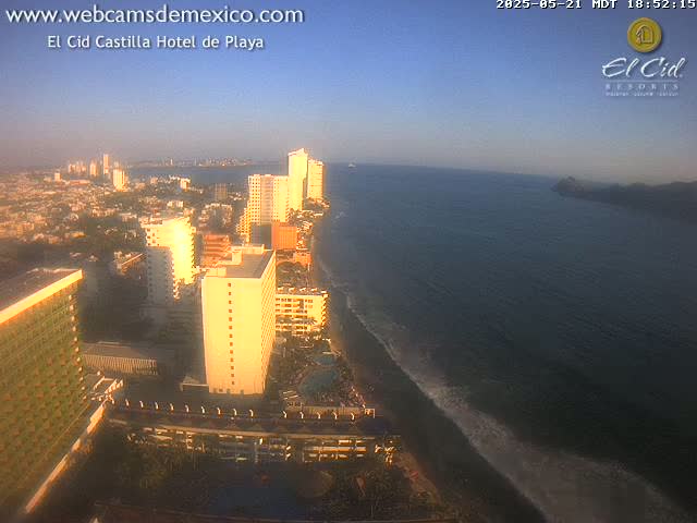 An aerial view of a coastal city at sunset, showing high-rise hotels, a beach with gentle waves, and calm ocean waters.