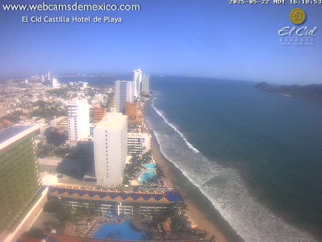 An aerial view shows a sandy beach with gentle waves lapping the shore, numerous high-rise hotels, and a clear, sunny sky.